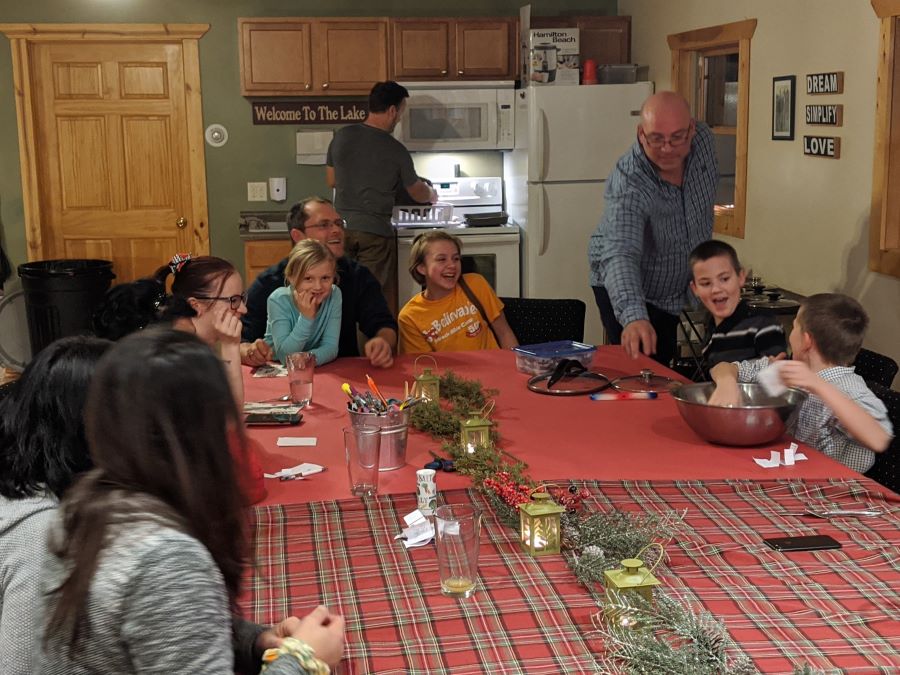 Family plays game at table decorated for Christmas while men cook and serve