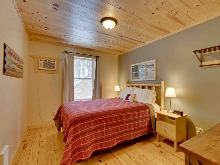 A red plaid quilt topped queen bed in bedroom with pine floor and ceiling of a lakeside cabin.
