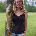 young women with blond wavy hair smiles while holding large walleye fish she caught