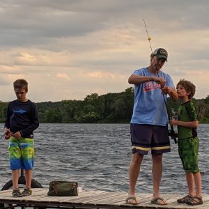A father and two sons stand on a dock fishing. The father is baiting one son's hook.