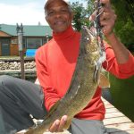 Older man with cap on kneels on a dock with a large walleye he caught