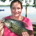 Smiling Woman holds up large black crappie in front of a lake.