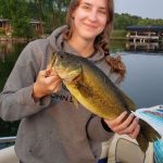 Young woman with brown hair in a braid holds a largemouth bass while on a boat on a lake