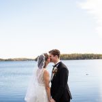 A bride and groom kiss next to a brilliant blue lake under blue sky