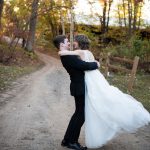 groom lifts bride off her feet while standing on a dirt road in the fall colored forest