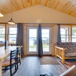 View of lake through glass doors and 2 windows in lakeside cabin. Pine table with chairs and log futon in the room