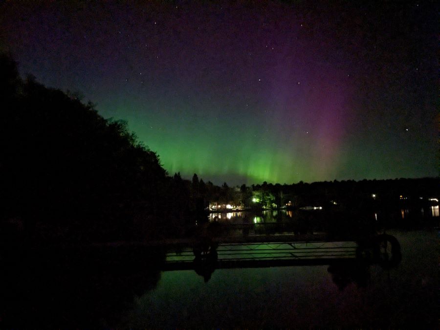 Beautiful northern lights with bright green and purple colors shine over a dark lake with a dock 