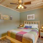 Lake cabin rental bedroom with queen bed with patchwork quilt and log pine headboard. Lamps on side tables and ceiling fan with lights on pine ceiling