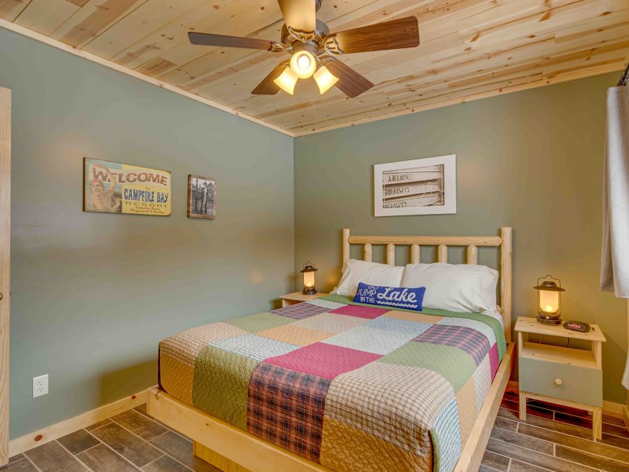 Lake cabin bedroom with Queen Bed with patchwork quilt and log pine headboard. Lamps on side tables and ceiling fan with lights on pine ceiling