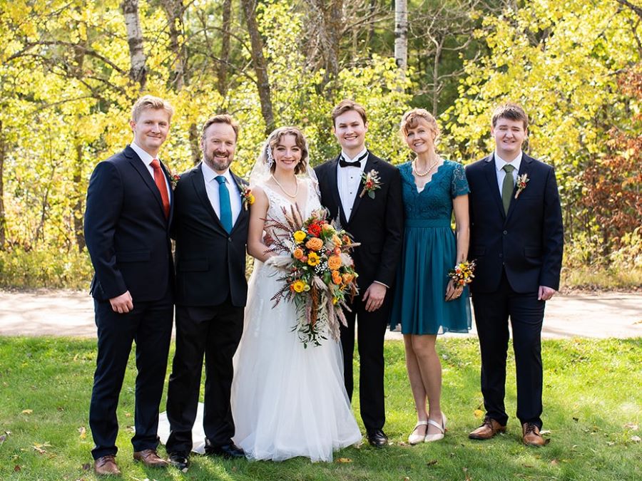 A family of 3 young men, a father with a beard, a mother with blond hair pulled up, and a young bride holding a bouquet stand in front of the woods on a beautiful fall day