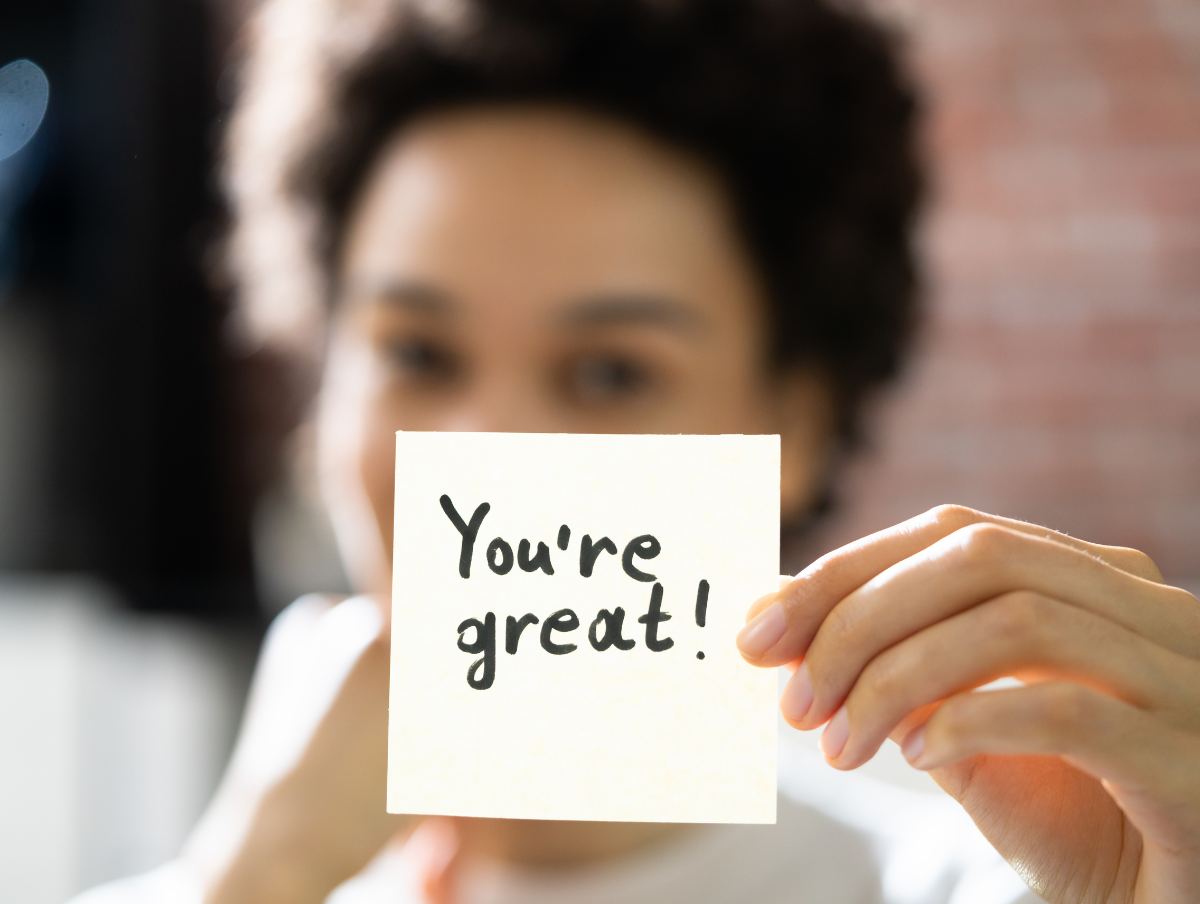 A woman holds up a sticky note that says, "You're great!", an example of words of affirmation
