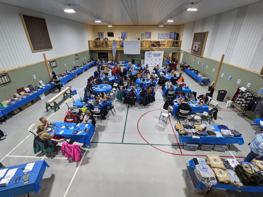 Several round and rectangular tables with bright blue tableclothes are in a gymnasium. People and products from Women Anglers of MN are gathered there.