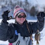Women with pink stocking cap and sunglasses holds fish caught while ice fishing on Fish Trap Lake