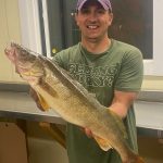 man with Vikings Football cap and green shirt holds a huge walleye while in a fish cleaning house