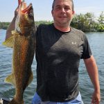 man in black shirt holds a large walleye vertically while on a boat
