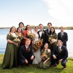 Five bridesmaids in olive silk dresses and groomsmen in navy suits with olive ties and the bride and groom stand and squat in front of a blue lake with blue sky