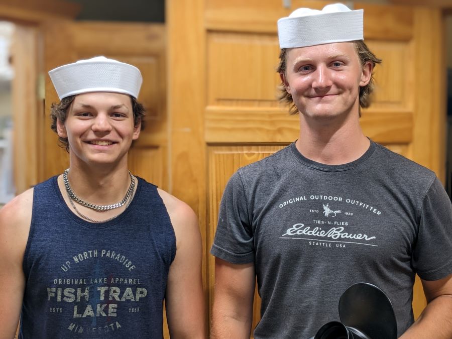 Two young men with tee shirts and white sailor hats on smile while one holds a boat propeller.