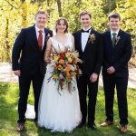 Bride, groom, and his two brothers stand in the grass in front of gorgeous fall colored trees