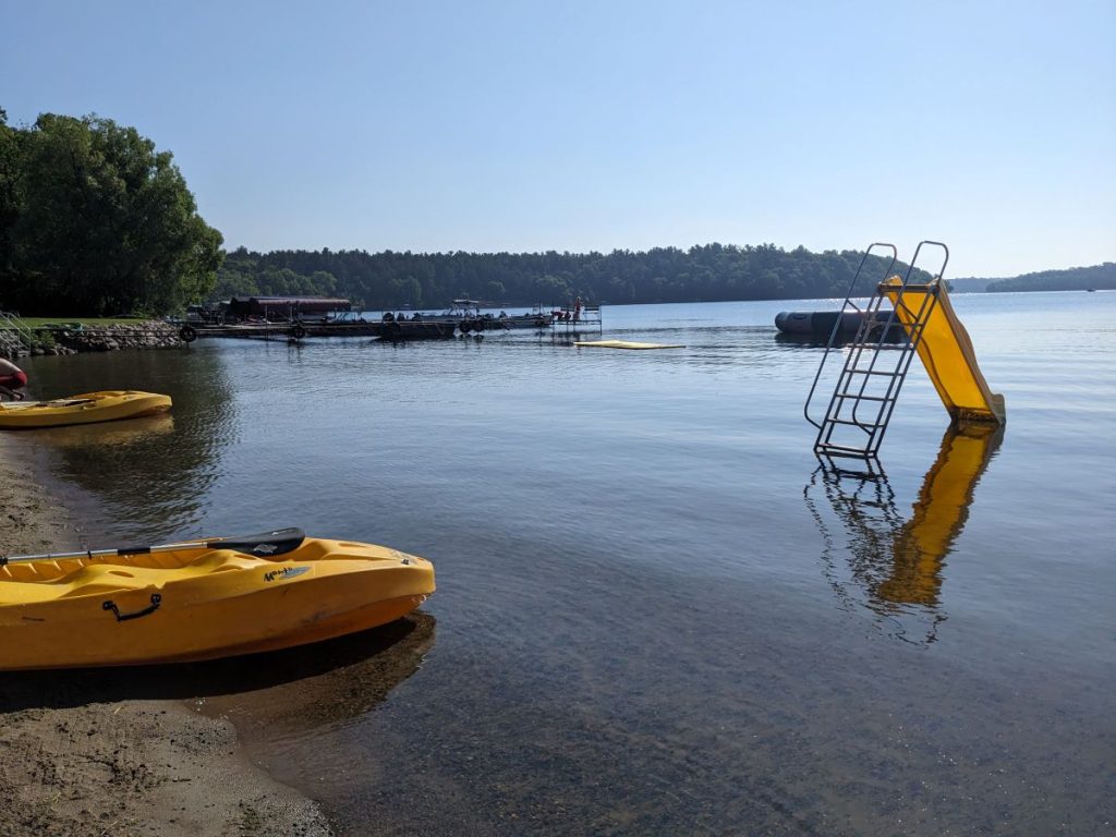 Beach with two yellow kayaks on the sand and view of small yellow slide, docks, and trampoline in the lake