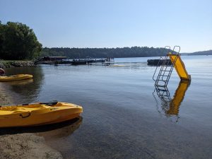 Beach with two yellow kayaks on the sand and view of small yellow slide, docks, and trampoline in the lake