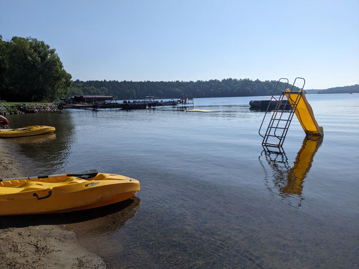 Beach with two yellow kayaks on the sand and view of small yellow slide, docks, and trampoline in the lake