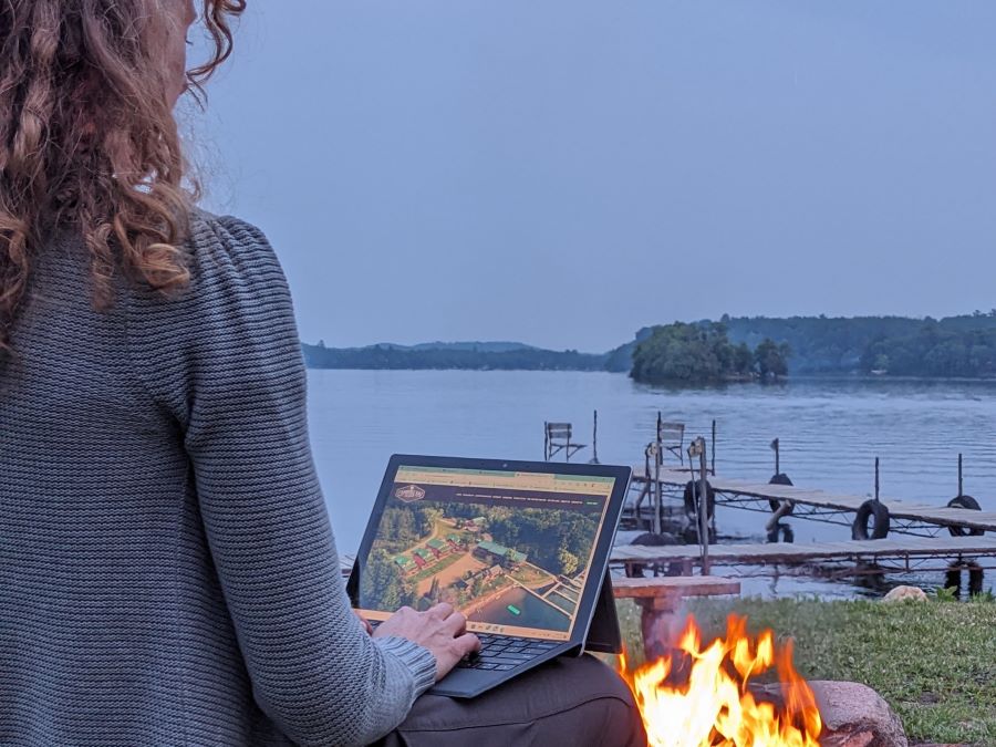 A woman works on her laptop outside by a campfire by the lake. Docks and an island are in the water