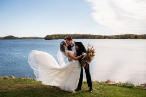Bride with dress blowing in the wind is being held by groom while kissing in front of Fish Trap Lake at a Minnesota resort