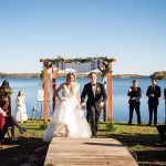 A bride and groom walk over a wooden bridge in front of a lake with a chuppah at the wedding recessional