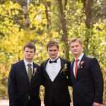 Two brothers in suits flank their groom brother while standing in the fall forest