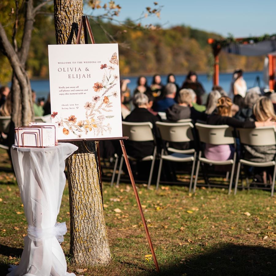 Wedding welcome sign on an easel and programs on a white tulle wrapped pedestal outside by people in chairs for the ceremony