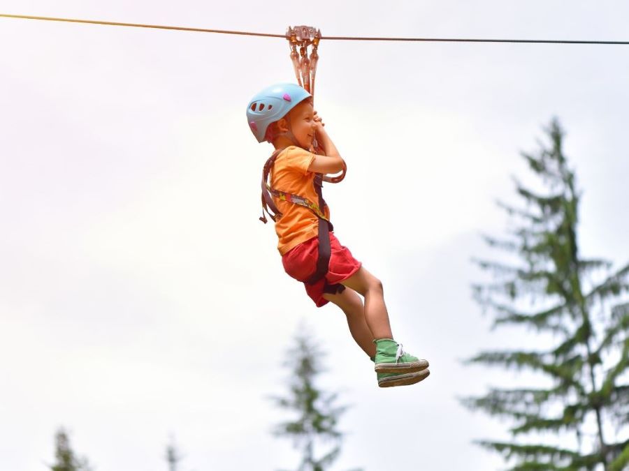A boy in a blue helmet and orange shirt ziplines in the sky with pine tree visible