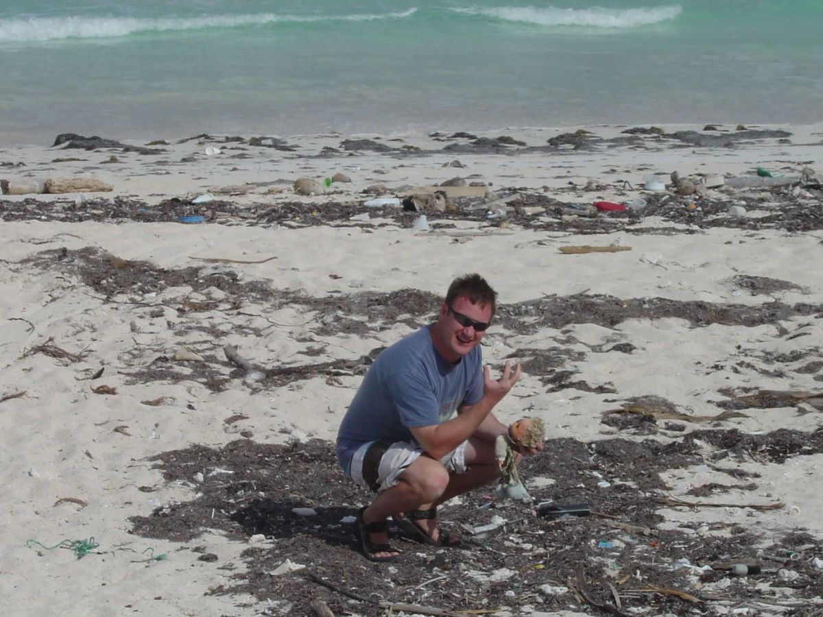 Man squats on polluted beach disgusted by the plastic and garbage. With specific goals to reduce single use plastic, maybe we can help.
