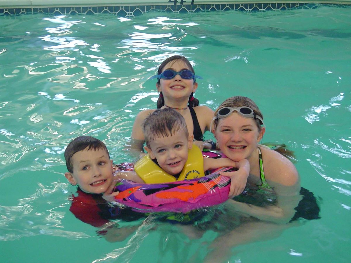 A young girl with swim goggles smiles as her sister and cousin hold onto a little boy in a floatie while at an indoor water park like one of the three in the Brainerd Lakes Area