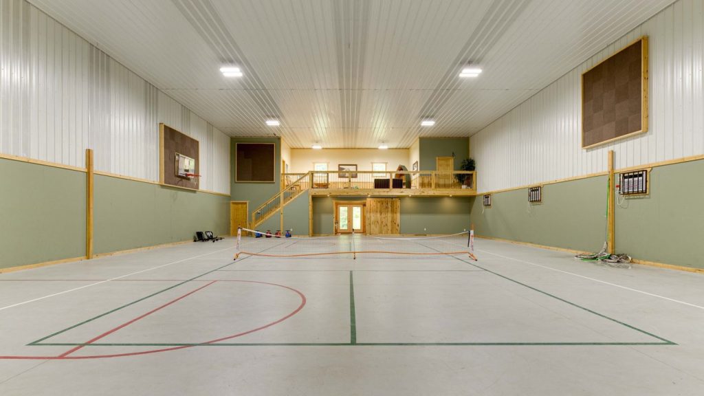 Gym floor showing basketball hoops and lines, pickleball net and floor lines, a volleyball net on the floor, and fitness room on the far end