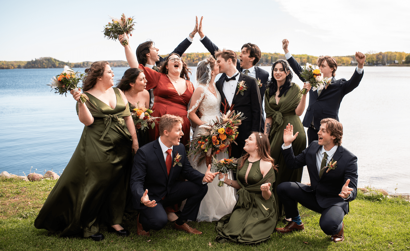 A group of four bridesmaids, one maid of honor, four groomsmen, one best man, and a bride and groom are gathered in front of a lake. Everyone is cheering while the bride and groom kiss.