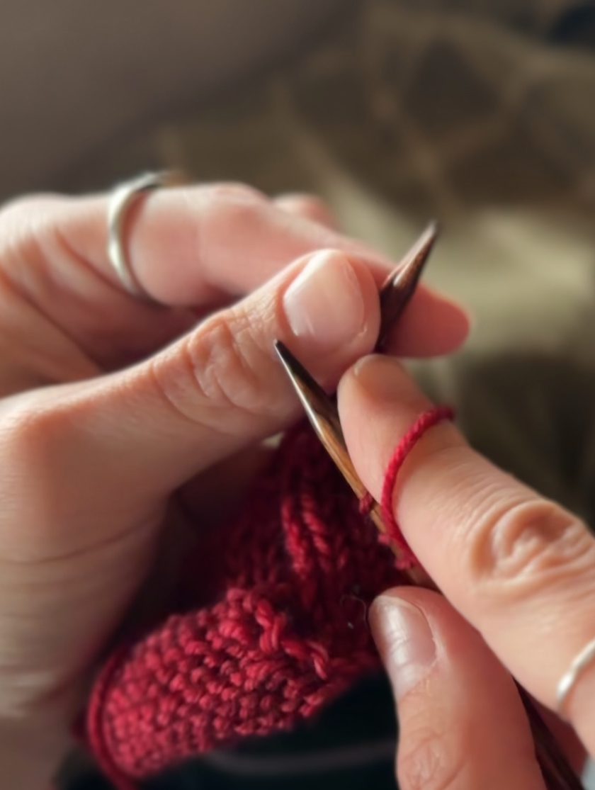 A close-up shot of fingers while knitting. The work is held between the hands on wooden needles, and a red thread is wrapped around the index finger on the knitter's right hand.