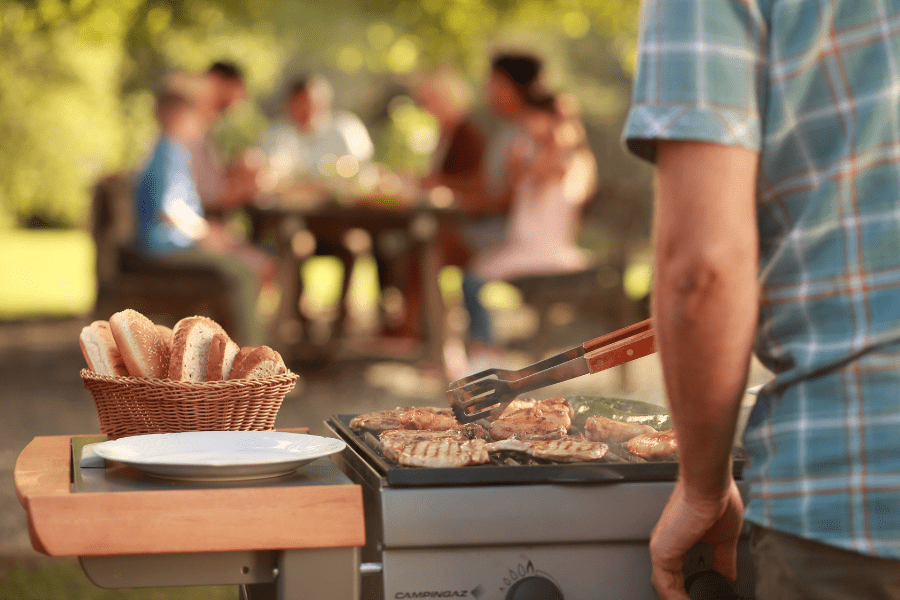 We see the arm and side of a man attending a grill, latent with burgers. Beside it sits a basket of buns. In the background, blurred, his family gathers for a meal.