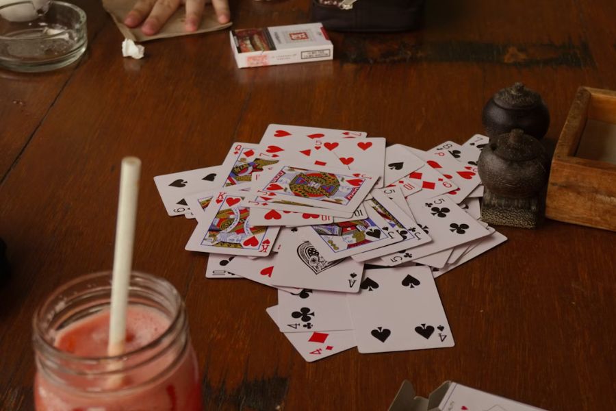 A pile of cards lay face-up on a wood table. Situated around it are cups and hands, as though this were the middle of a card game.