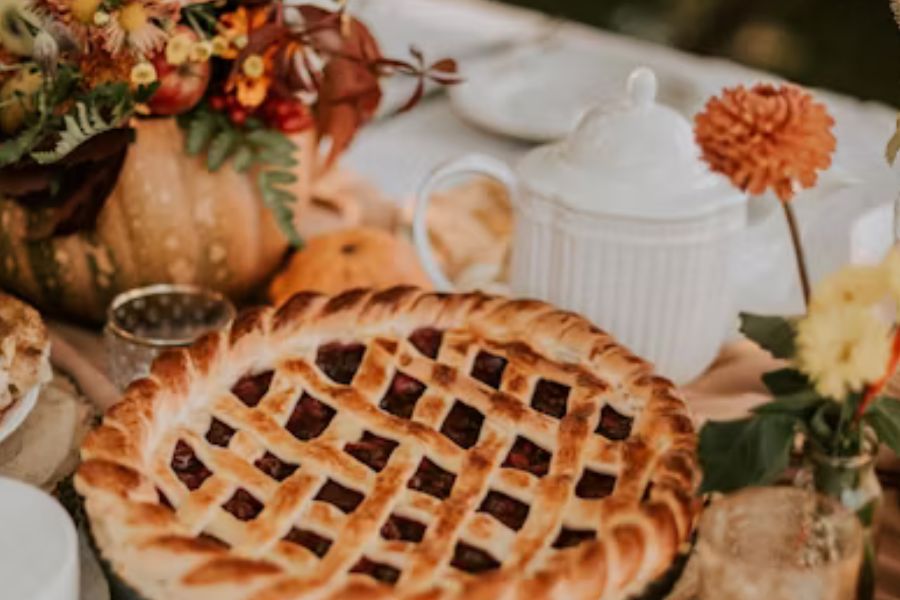 A large pie, likely cherry, with latticed top that shows the filling. Around it are autumn grasses and flowers in vases.