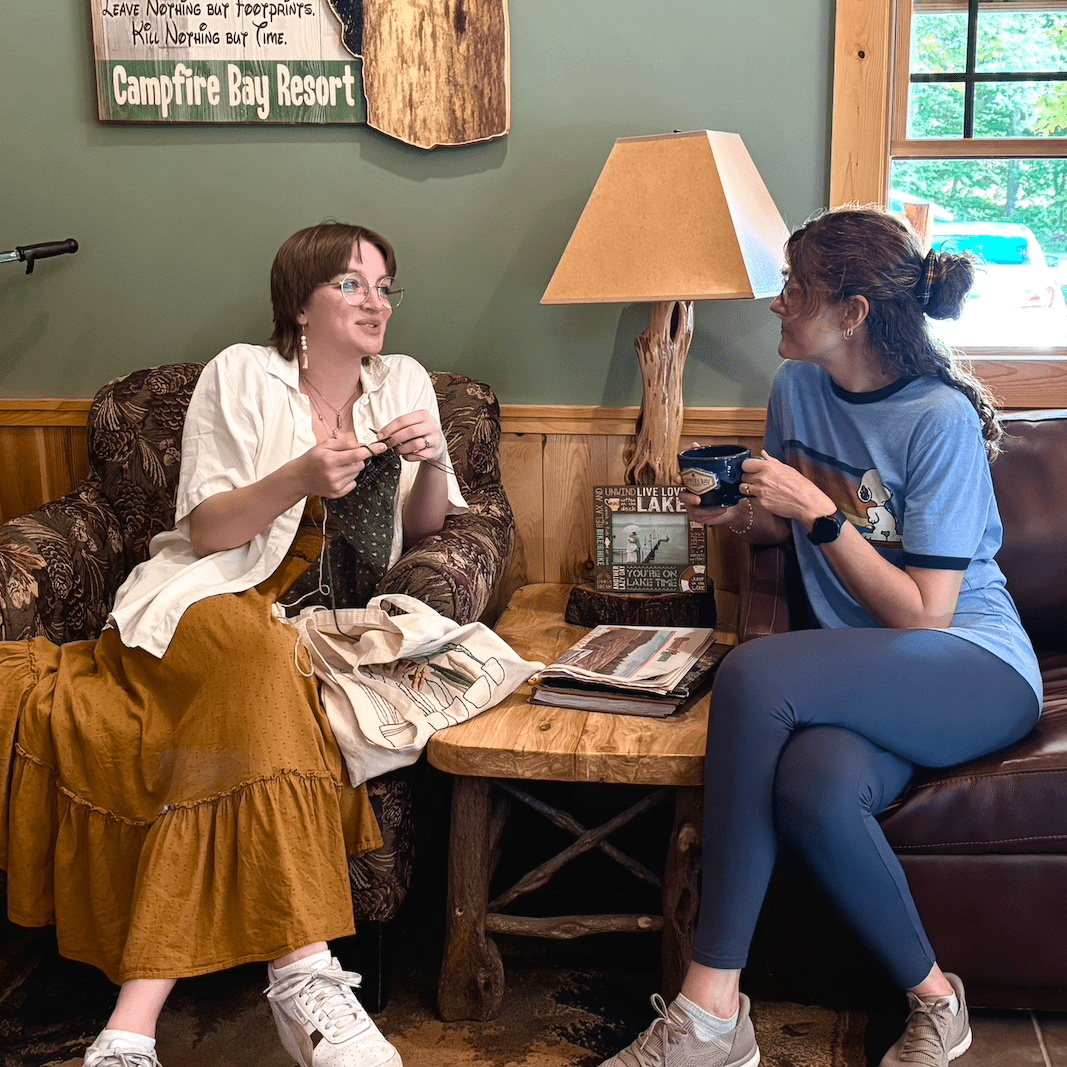 Two women sit in armchairs. They lean towards each other. One is knitting and talking, the other is sipping coffee.