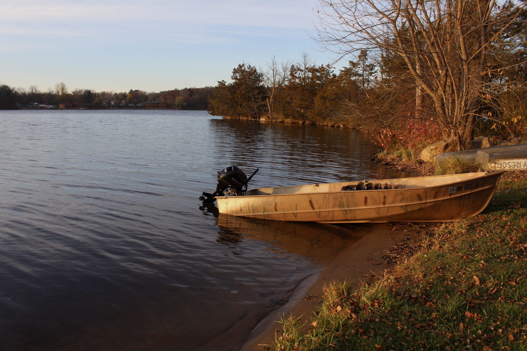 Image of a boat on the bay of an island. There are fallen leaves around it and Campfire Bay Resort can be seen in the distance across the lake. 