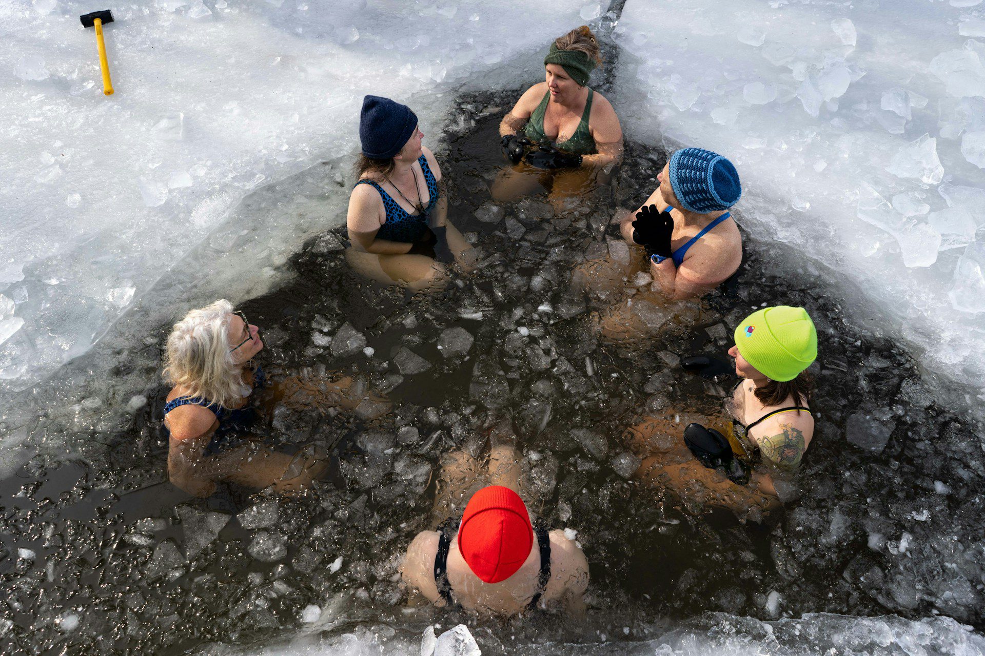 Six women with hats and gloves cold plunging in a lake with ice