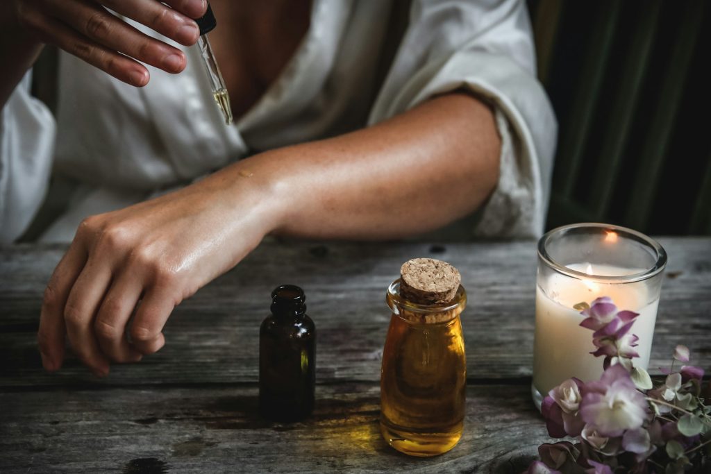 Image of a women pouring oil on her arm. A bottle of essential oil, a jar of oil, and a lit candle can be seen