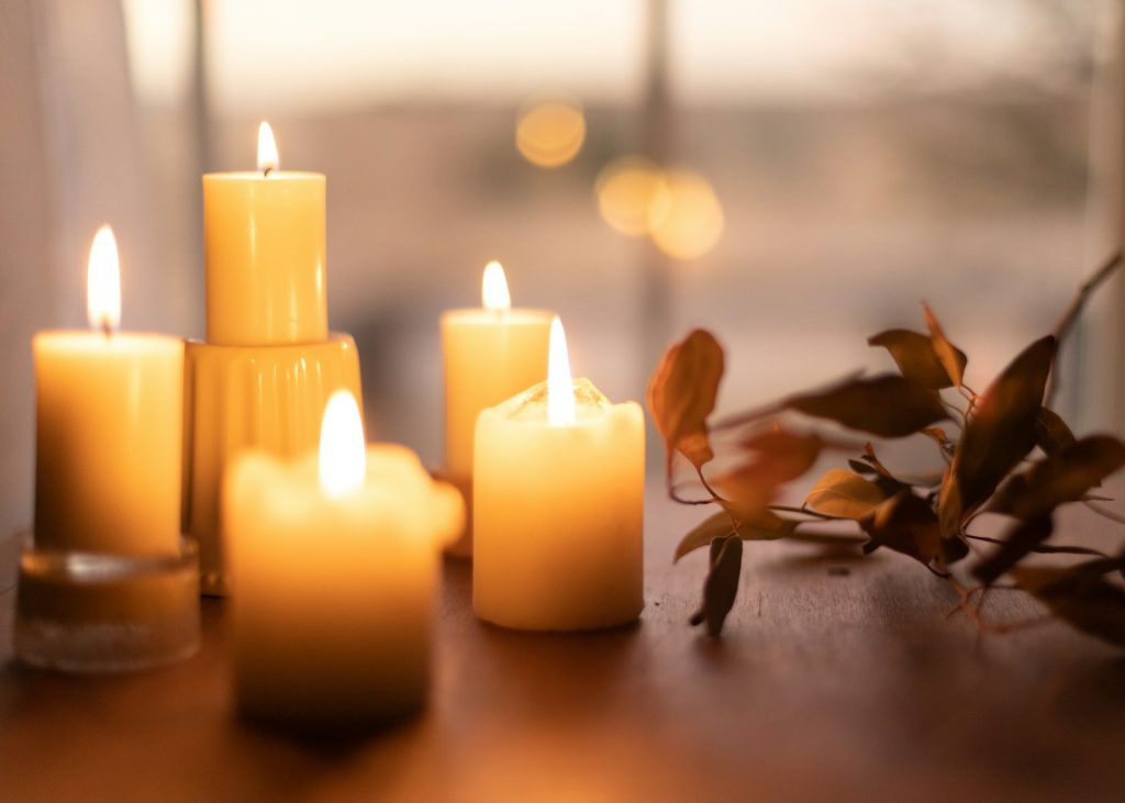 Image of white pillar candles glowing while lit, dried leaves can be seen next to them