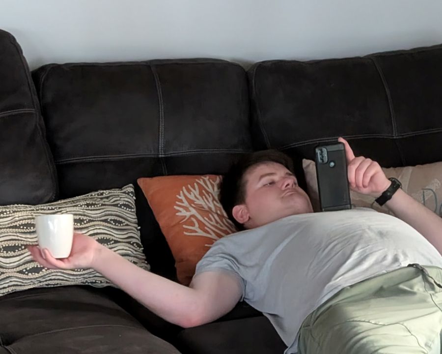 A man looks at his phone while lying down on his back and holding a coffee cup to the side