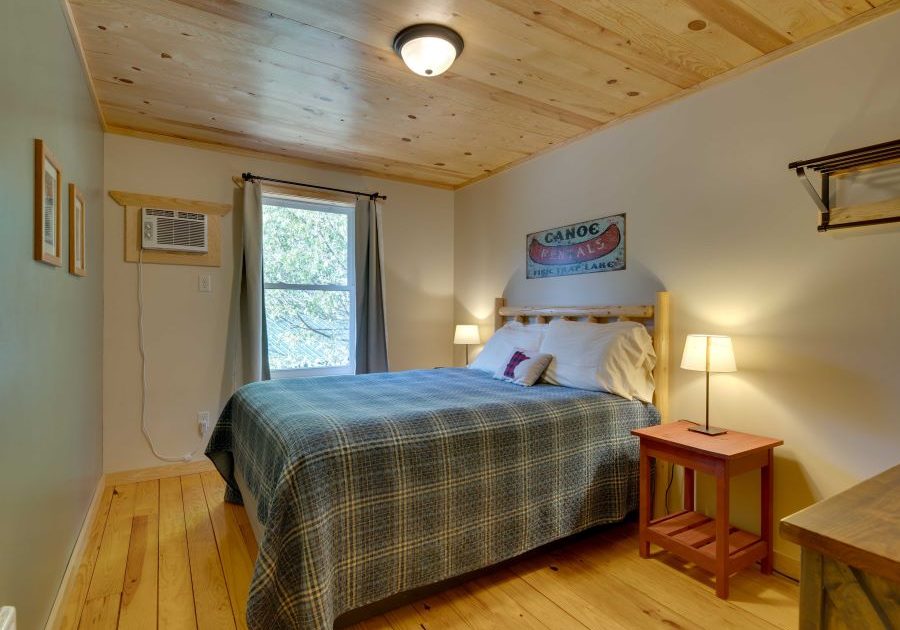 Cabin bedroom with pine floor, ceiling, and headboard of queen bed with blue plaid quilt