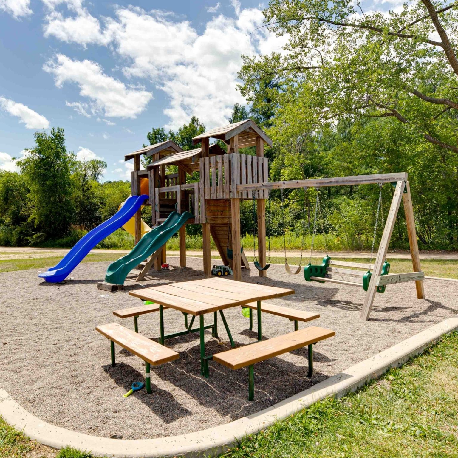 Playground with a swingset and elevated playhouse with 2 slides on a pea gravel concrete rimmed pad on a sunny day