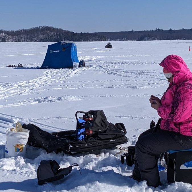 Fisherman in pink jacket sitting on a snow covered lake fishing near a blue portable fish house